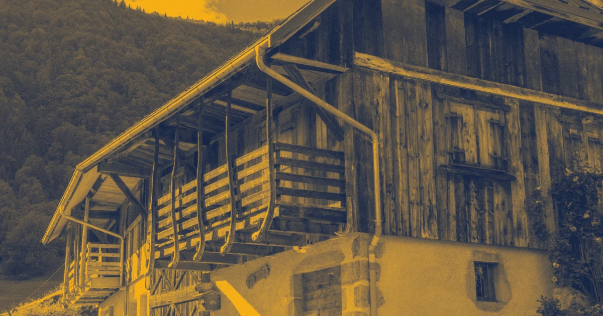Ancienne ferme en bois sur un rez-de-chaussée maçonné, avec un balcon en bois sous l'avancée du toit, devant un versant boisé.