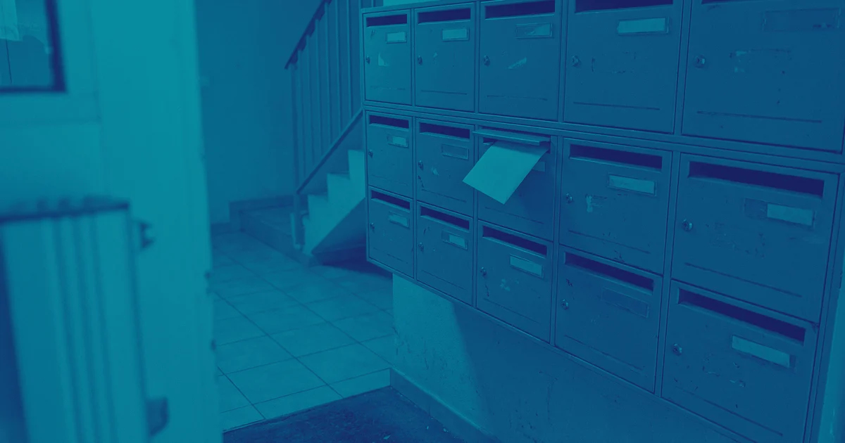 Boîtes aux lettres métalliques alignées dans le hall d'un immeuble, porte-noms vierges, une enveloppe dépasse de l'une d'elles ; escalier au fond.